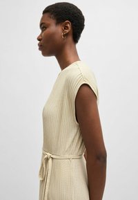 Light beige ribbed dress with short sleeves, a fitted silhouette, and a tied waist detail, featuring a smooth texture and elegant design.