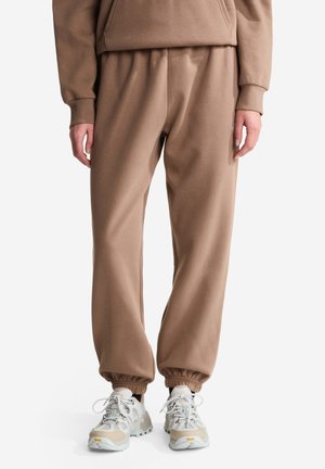 Brune joggerbukser med elastiske manchetter, båret med beige og hvide snøresneakers, vist fra midt på taljen til fødderne på hvid baggrund.