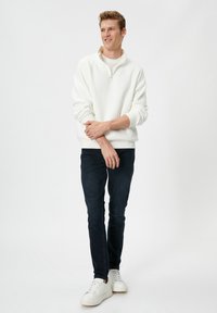 Jonge man staat met gekruiste armen, draagt een witte sweatshirt met kwartrits, donkere jeans en witte sneakers op een effen lichte achtergrond.