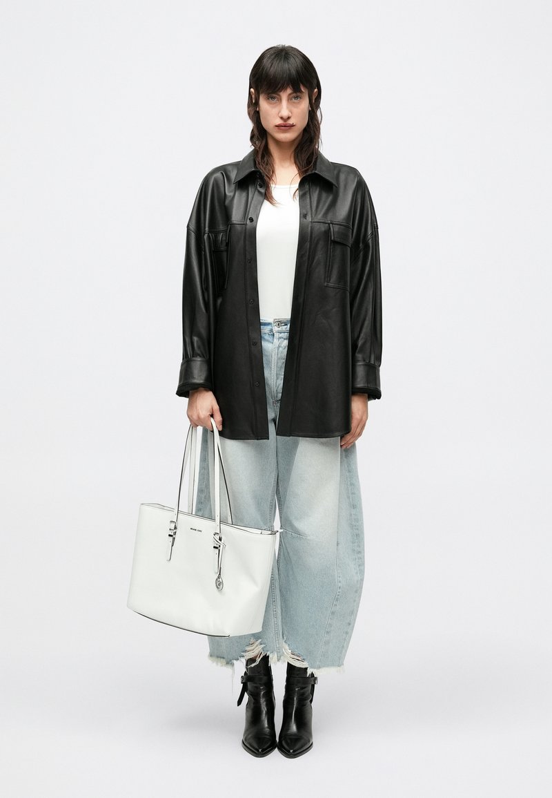 Camisa casaco oversized em pele preta, top branco, jeans azul claro desgastado de perna larga, botas pretas até ao tornozelo e uma bolsa tote branca com detalhes prateados.