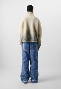 Kunstfelljacke in Beige mit verblasstem Grau, kombiniert mit übergroßen blauen Jeans aus Denim mit ausgefransten Säumen und weißen Farbspritzern.