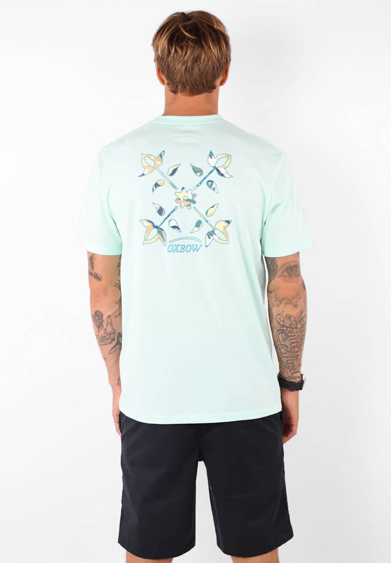 T-shirt en coton vert menthe avec un graphique floral coloré au dos ; manches courtes et coupe décontractée. Porté avec un short noir.