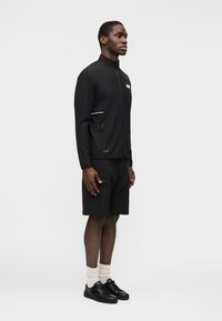 Άνδρας όρθιος, φοράει μαύρο μπουφάν με φερμουάρ, μαύρα σορτς με τσέπη, λευκές κάλτσες και μαύρα αθλητικά παπούτσια, μπροστά σε απλό φωτεινό φόντο.