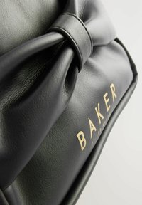Crna kožna torbica s istaknutim detaljem mašne i zlatnim natpisom "BAKER" na prednjoj strani. Glatka tekstura.