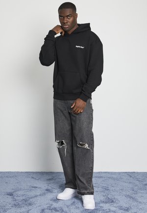 Svart hettegenser med frontlomme, med hvit logo tekst. Kombinert med slitte grå jeans og hvite joggesko på en teksturert blå overflate.