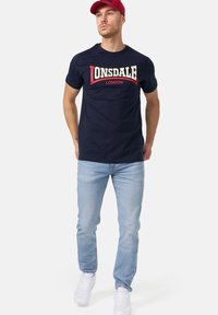 Navy bomulds t-shirt med hvid og rød "LONSDALE LONDON" logo, parret med lyseblå jeans og hvide sneakers. Rød kasket fuldender looket.
