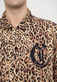 Camisa de manga corta con estampado de leopardo y diseño de botones. Presenta botones negros y un gran logo de letra negra en la parte frontal.