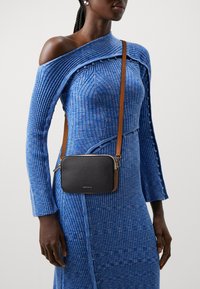 Borsa a tracolla nera in pelle con superficie texturizzata, hardware dorato e tracolla marrone, indossata sopra un vestito in maglia blu a coste.