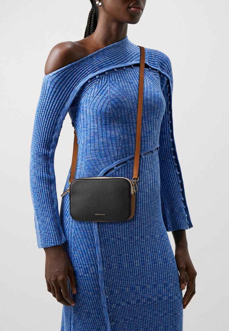 Borsa a tracolla nera in pelle con superficie texturizzata, hardware dorato e tracolla marrone, indossata sopra un vestito in maglia blu a coste.