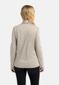 Donna con capelli biondi che indossa una maglietta a maniche lunghe grigia chiara e pantaloni neri, vista di spalle su uno sfondo bianco semplice.