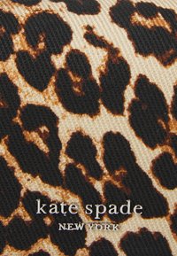 Un tissu à motif léopard présente des taches noires et brunes sur un fond beige clair. Le texte "kate spade NEW YORK" en argent est positionné au centre.