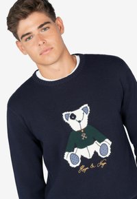 Suéter de punto azul marino con un motivo de oso en tonos azules. El oso lleva un suéter verde, y la bordado en dorado dice "Harper & Meyer."