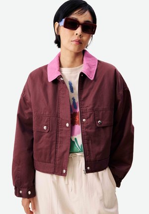 Femme portant une veste maroon surdimensionnée avec un col rose, une chemise à motifs en dessous, un pantalon crème et des lunettes de soleil rectangulaires foncées.