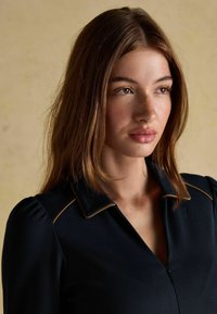 Must must esip, millel on lukuklapp, krae ja paisutatud varrukad. Krae ääres on kuldne ääris. Sile tekstuur.