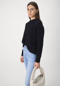 Schwarzer Strickpullover mit hohem Ausschnitt, kombiniert mit hellblauen High-Waist-Jeans mit Knopfakzenten und einer cremefarbenen Schultertasche.