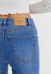 Vaqueros de denim azul con un parche de cuero etiquetado "PULL&BEAR", mostrando bolsillos traseros y detalles de costura, diseño ajustado y textura de lavado ligero.