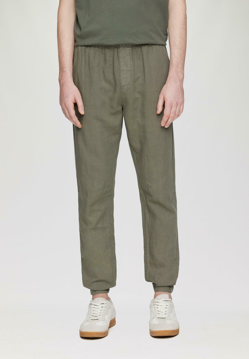 Spodnie w oliwkowym kolorze z elastycznym paskiem w talii i zwężanymi nogawkami, wykonane z lekkiego lnu, z delikatnymi detalami szwów, połączone z białymi trampkami.