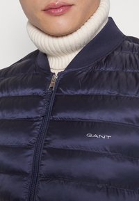 Giubbotto imbottito blu navy con cuciture orizzontali e chiusura a zip; presenta un collo a coste blu navy e un logo argento sul lato sinistro.