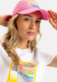 Gorra de algodón rosa con visera curva, que presenta el texto "BYRON BAY" y gráficos de sol. Llevada con una camiseta blanca que muestra un estampado tropical colorido.