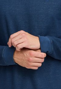 Camicia a maniche lunghe di colore blu navy con texture e polsini a coste. Mani incrociate attorno al polso, evidenziando il design aderente della camicia e il tessuto liscio.
