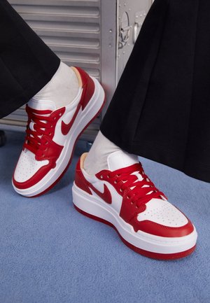 Persona con zapatillas blancas y rojas con cordones rojos y pantalones negros, sentada en un suelo alfombrado azul claro junto a un objeto metálico.