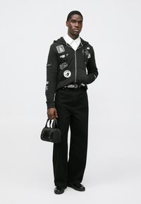 Juodas džemperis su gausiais pleistrais, derinamas su juodais aukšto liemens, platėjančiais džinsais. Aksesuarai: maža juoda rankinė ir juodi batai.