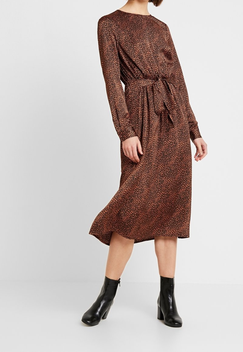 Femme portant une robe midi marron et noire tachetée, manches longues et ceinture à la taille, associée à des bottines noires.