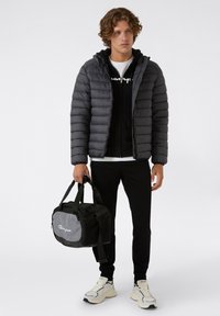 Kurtka puchowa w ciemnoszarym kolorze, na zamek błyskawiczny, z kapturem; czarna bluza pod nią; czarne spodnie; niesie czarną torbę sportową z szarym akcentem.
