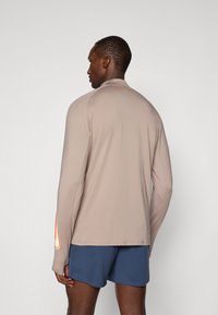 Camară sport cu mânecă lungă, de culoare bej, cu o textură netedă, având un mic logo aproape de gât și accente colorate pe mâneci.