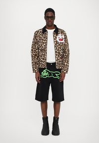 Giacca bomber con stampa leopardata, colletto nero, t-shirt bianca, pantaloni corti neri con grafica verde neon e stivali neri alla caviglia.