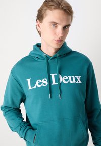 Turquoise hoodie with white "Les Deux" text. Features a front pocket, drawstrings, and ribbed cuffs. Fabric texture appears soft.