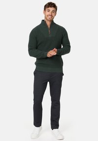 Suéter sólido de canalé verde oscuro con media cremallera, que presenta un patrón de tejido texturizado. Combinado con pantalones oscuros ajustados y zapatillas blancas.