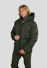 Homme aux cheveux blonds mi-longs portant une veste matelassée verte foncée zippée et un pantalon cargo assorti, sur un fond clair uni.