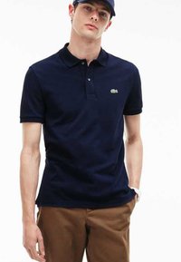 Polo de color azul marino hecho de algodón, con cuello, dos botones y un pequeño logo verde en el lado izquierdo del pecho. Combinado con pantalones marrones.
