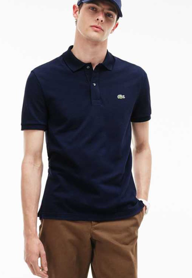 Polo de color azul marino hecho de algodón, con cuello, dos botones y un pequeño logo verde en el lado izquierdo del pecho. Combinado con pantalones marrones.