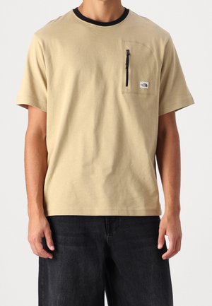 Beżowa koszulka z krótkim rękawem i okrągłym dekoltem z czarnym akcentem. Posiada kieszeń na piersi po lewej stronie zapinaną na zamek oraz naszywkę z logo. Miękka w dotyku.