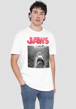 Young man wearing a white T-shirt with the red "JAWS" logo and a shark rising toward a swimmer on the front, paired with blue jeans.