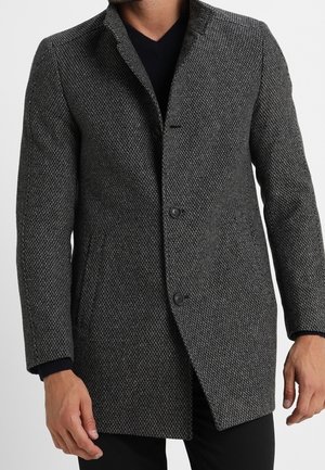 Homme portant un manteau texturé gris foncé boutonné par-dessus un pull noir, les mains détendues le long du corps sur un fond neutre.