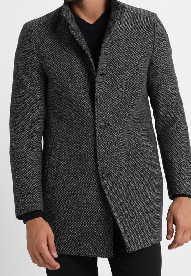 Homme portant un manteau texturé gris foncé boutonné par-dessus un pull noir, les mains détendues le long du corps sur un fond neutre.