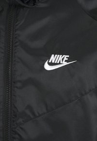 Černá nylonová bunda s plným zipem, prošívanou texturou a výrazným bílým vyšitým logem Nike na hrudi.