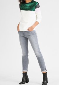 Pulóver con parte superior de lentejuelas verdes y sección inferior blanca. Jeans ajustados grises, con puños doblados y botas negras hasta el tobillo. Textura suave, estilo casual.