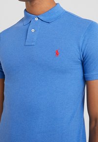 Blå polotröja i bomull, med klassisk krage, tvåknäpps placket och en liten röd logotyp på vänster bröst.