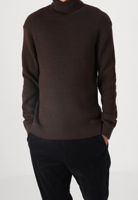 Brązowy sweter z wysokim kołnierzem, wykonany na drutach, o fakturowanym wzorze, z długimi rękawami i luźnym krojem, zestawiony z ciemnymi spodniami.
