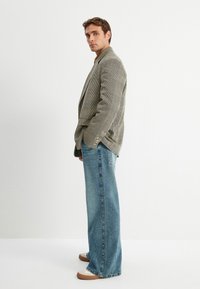 Blazer a cuadros en tonos apagados, combinado con jeans de denim azul de pierna ancha. Los zapatos presentan un diseño en beige y gris. Fondo limpio.