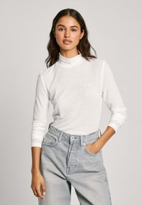 Haut à col roulé blanc à manches longues avec un détail de logo subtil, associé à un jean taille haute gris clair. Texture douce et coupe décontractée.