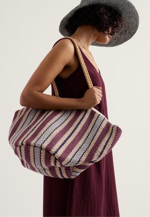 Frau trägt ein ärmelloses, bordeauxfarbenes Kleid und einen breitkrempigen Hut. Sie hat eine große gestreifte gewebte Tragetasche mit Seilgriffen über der Schulter.