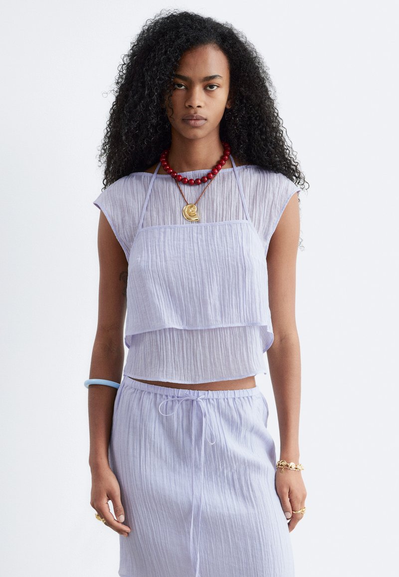 Femme aux longs cheveux bouclés portant un haut sans manches violet clair en couches et un pantalon assorti, un collier de perles rouges et des bijoux en or sur fond blanc.