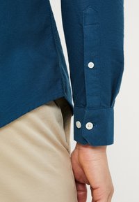 Niebieska koszula z długim rękawem z zapięciem na guzik i wyprofilowanym mankietem, uszyta z teksturowanej tkaniny, w połączeniu z jasnobeżowymi spodniami.