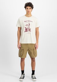 T-shirt di cotone crema con un design grafico rosso che raffigura una donna che tiene del cibo, abbinata a pantaloni cargo color khaki e sneakers nere.
