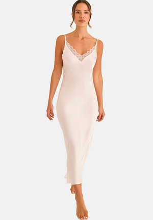 Vrouw draagt een lange, witte slipjurk met kanten randjes op de V-hals, loopt op blote voeten tegen een eenvoudige achtergrond.